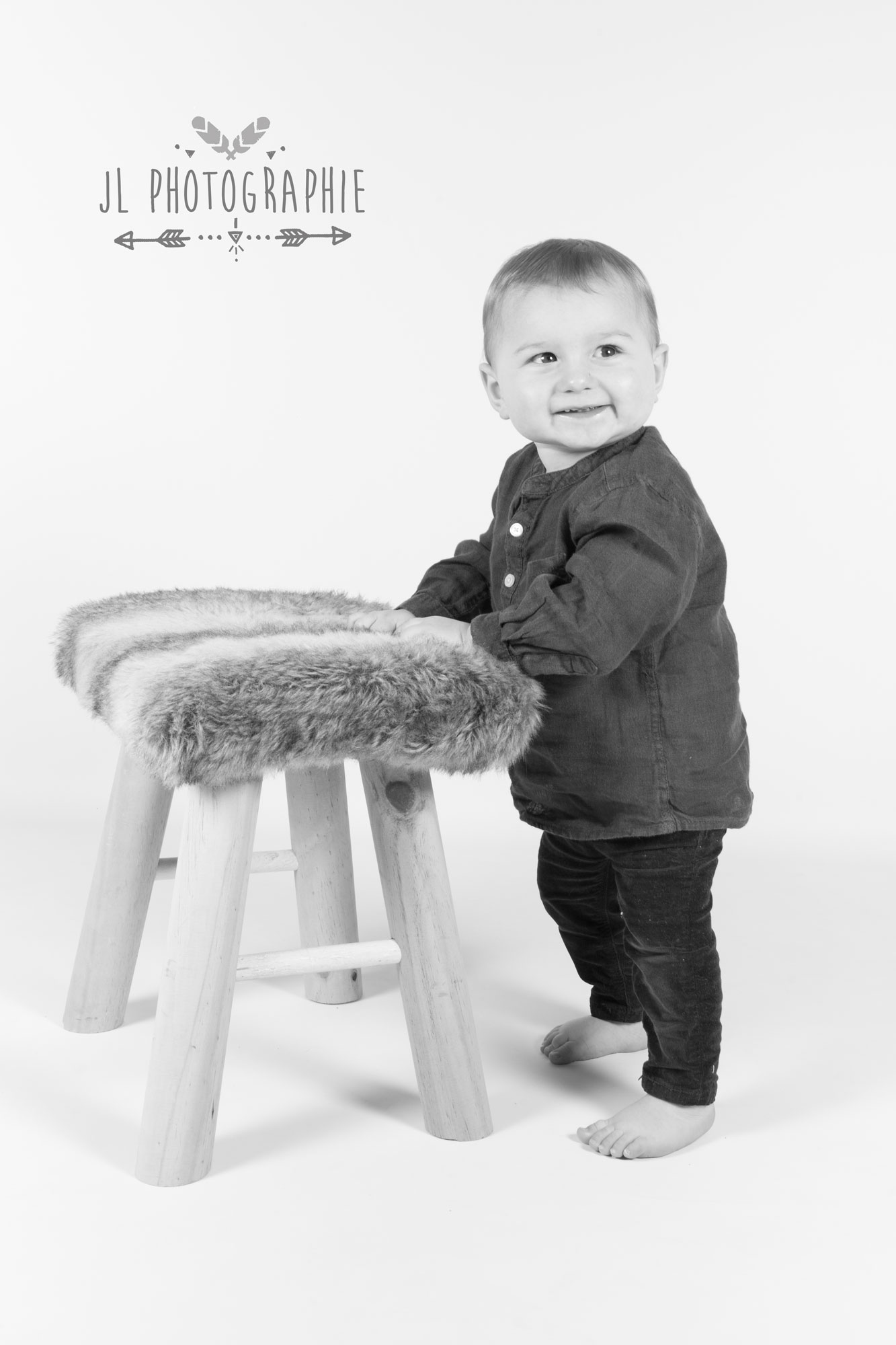 Photographie en noir et blanc d'un enfant debout devant un tabouret