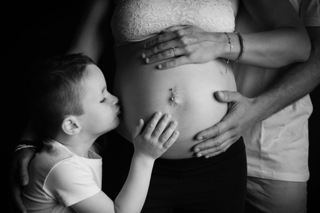 Photographie de grossesse en noir et blanc avec un enfant qui fait un bisou sur le ventre
