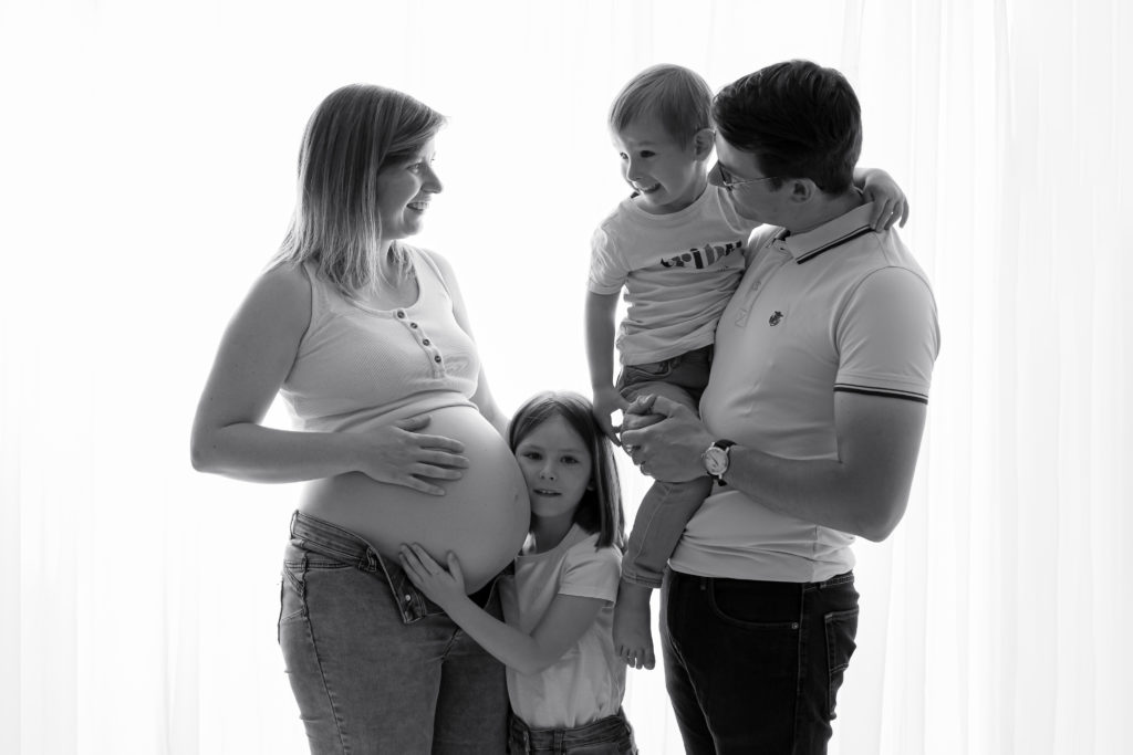 Séance photo grossesse en studio femme enceinte devant une fenêtre en noir et blanc et en famille