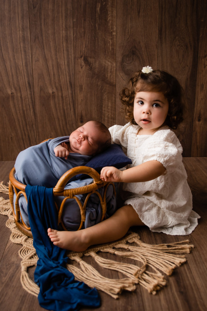 Séance photo nouveau né garçon et de sa soeur en studio dans l’Oise positionnée dans un panier couleur bleu